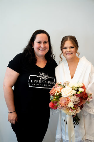 Image of Niki, the owner of Pepper & Fern, posing with a bride on her wedding day. Niki is in her Pepper and Fern logo shirt anf the bride is in her getting ready robe holding her just delivered wedding flowers