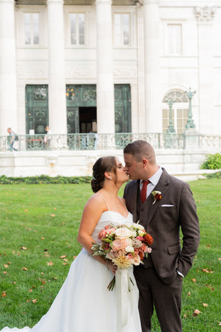 Image of bride and groom in front of Newport mansion. Bride is holding an early fall bridal bouquet with muted colors including terracota.