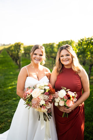 Image of bride with her bridesmaid. Bridesmaid is in a rust colored bridesmaid dress. Both are holdign floral bouquets infront of vineyard.