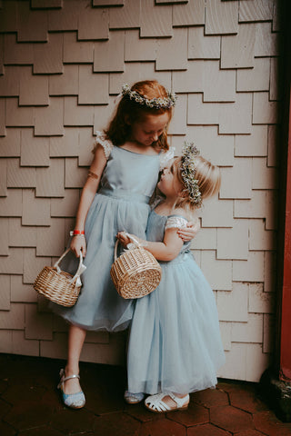 image of two flower girls in pale blue dresses. The smallest one is looking up to the bigger one with her looking down. They are wearing pale blue dresses and floral crowns, carrying wicker baskets.