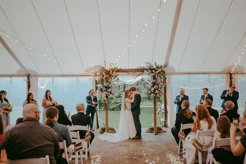 Image of couple under an arch during their wedding ceremony during the bride and groom's firrst kiss. The wedding arch has an elegant floral dsign on either side that make a statement.