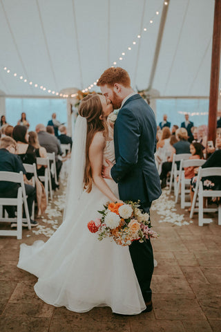 Image of bride and groom walking away from their wedding ceremony, stopped at the end of the aisle for a kiss. The bride is holding a whimsical bouquet with wildflowers with a summer color palette with pale oranges and pink.