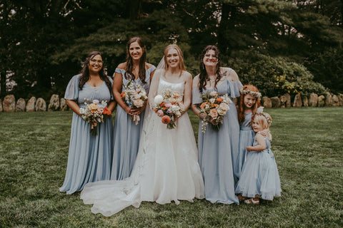 Image of bride with her bridal party. Bride is in center with bridesmaids and flower girls on either side. Bridesmaids and flower girls are in pale blue dresses. Flower girls are wearing flower crowns with bridesmaids carriyng wildflower bouquets.