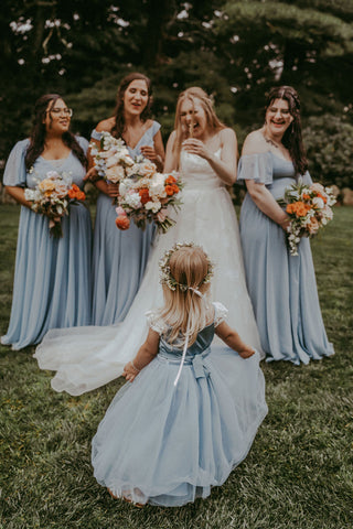 Image of bride with bridesmaids all happily smiling and laughing. Shows the back of the flower girl walking towards them. Assumingly causing them to smile and laugh.