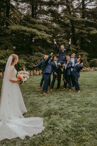 Image of bride holding wildflower bright bouquet looking back at the groom being held up by the groomsmen.