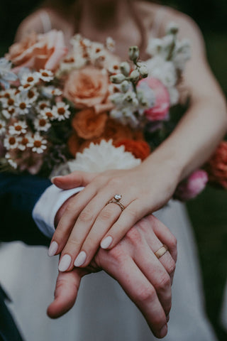 Image of bride and groom's hands showing off their wedding bands in front of the bride's bridal bouquet with wildflowers, shades of pale oranges and pinks.