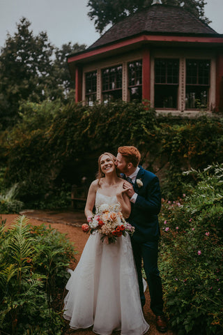 Image of bride and groom. They are in the gardens at the Estate at Moraine Farm. The groom is kissing the bride's cheek with the bride having a big smile on her face.