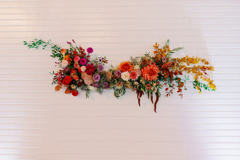 Image of floral installation with muted fall colors, flowers include dahlias, marigolds, sedum, celosia, ranunculus, lisianthus, mums, leucadendron, asclepia, eucalyptus, and amaranthus.