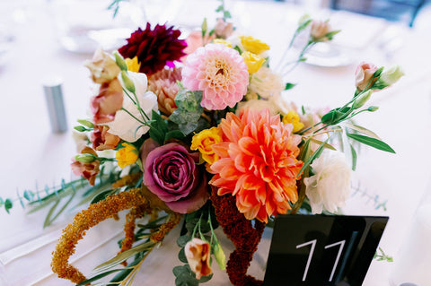 Image of fall wedding centerpiece. Image is a close up of the flower arrangement including dahlias, marigolds, sedum, celosia, ranunculus, lisianthus, mums, leucadendron, asclepia, eucalyptus, and amaranthus. It has muted fall colors including shades of pink, purple, orange, yellow and white.
