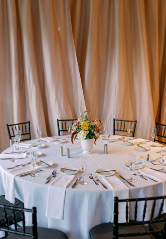 Image of wedding reception table set up at the Cape Club of Sharon. The image shows the beige fabric wall behind the table and the flower centerpiece with table settigns set up.