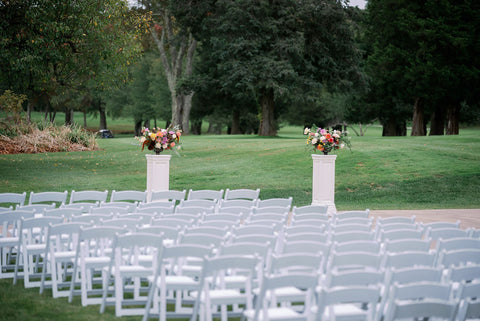 Image of the outdoor ceremony set up at the Cape Club in Sharon, a Massachusetts wedding venue. The image shows white garden chairs set up on the outdoor lawn space. There are two pedestals where the bride and groom with be with stunning floral designs.