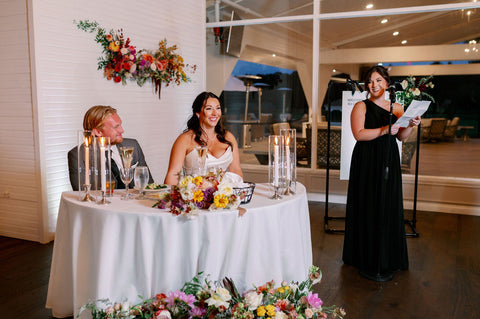 Image of bride and groom sitting at sweetherat table at the Cape Club of Sharon, a Masachusetts wedding venue. The sweetheart table is surrounded by a floral installation on the wall behding the couple, their ceremony flowers reused to add more color and some flowers on the tabel. Flowers include dahlias, marigolds, sedum, celosia, ranunculus, lisianthus, mums, leucadendron, asclepia, eucalyptus, and amaranthus in muted fall colors. The couple is laughing at the maid of honor speech. Yuo can see the maid of honor standing next to the couple reading her speech.