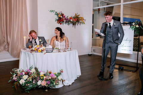 Image of best man giving wedding speech as the bride and groom laugh.