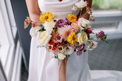 Zoomed in photo of a bridal bouquet for a fall wedding in muted fall colors. The bouquet is being held by the bride. The colors pop against the bride's sating white dress. The flowers include dahlias, marigolds, sedum, celosia, ranunculus, lisianthus, mums, leucadendron, asclepia, eucalyptus, and amaranthus.
