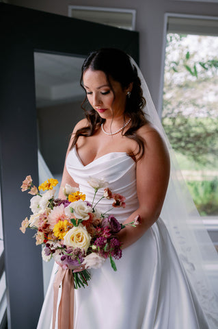 Image of bride holding and looking down at her bridal bouquet which has a muted fall color palette and Image of wedding tablescape at the Cape Club of Sahron, a Massachusetts wedding venue. The image highlgihts the wedding flower centerpieces which include muted fall colors and Zoomed in photo of a bridal bouquet for a fall wedding in muted fall colors. The bouquet is being held by the bride. The colors pop against the bride's sating white dress. The flowers include dahlias, marigolds, sedum, celosia, ranunculus, lisianthus, mums, leucadendron, asclepia, eucalyptus, and amaranthus.