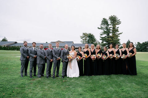 Image of wedding party lined up for a posed shot. The groomsmen are on side of groom all wearing gray suits. The bridesmaids are on the side of the bride, all wearing black dresses.