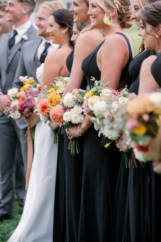 Image of zoomed in wedding party portrait from an angle. The zoome focuses on the brdiesmaids bouquets all lined up. The bouquets featured muted fall colors and flowers including dahlias, marigolds, sedum, celosia, ranunculus, lisianthus, mums, leucadendron, asclepia, eucalyptus, and amaranthus. The colors pop against the black bridesmaids gowns.