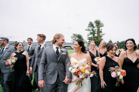 Image of bride and groom surrounded by their wedding party. Everyone is laughing and smiling. The bride and couple are looking at each other. The bride and bridesmaids are all holding bouquets in a muted fall colr palette.