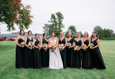 Image of bride and bridal party all lined up. The bride is standing in the middle surrounded by her bridesmaids who are wearing long black dresses. They are al holding bouquets in muted fall color palette including Image of couple portraits for a wedding couple at the Cape Club of Sharon, a Massachusetts wedding venue. The couple is standing on a road next to a tree. The bride is holding her bridal bouquet in a muted fall color palette including Image of wedding tablescape at the Cape Club of Sahron, a Massachusetts wedding venue. The image highlgihts the wedding flower centerpieces which include muted fall colors and Zoomed in photo of a bridal bouquet for a fall wedding in muted fall colors. The bouquet is being held by the bride. The colors pop against the bride's sating white dress. The flowers include dahlias, marigolds, sedum, celosia, ranunculus, lisianthus, mums, leucadendron, asclepia, eucalyptus, and amaranthus..