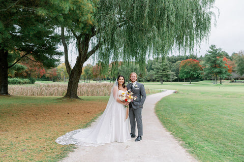 Image of couple portraits for a wedding couple at the Cape Club of Sharon, a Massachusetts wedding venue. The couple is standing on a road next to a tree. The bride is holding her bridal bouquet in a muted fall color palette including Image of wedding tablescape at the Cape Club of Sahron, a Massachusetts wedding venue. The image highlgihts the wedding flower centerpieces which include muted fall colors and Zoomed in photo of a bridal bouquet for a fall wedding in muted fall colors. The bouquet is being held by the bride. The colors pop against the bride's sating white dress. The flowers include dahlias, marigolds, sedum, celosia, ranunculus, lisianthus, mums, leucadendron, asclepia, eucalyptus, and amaranthus.