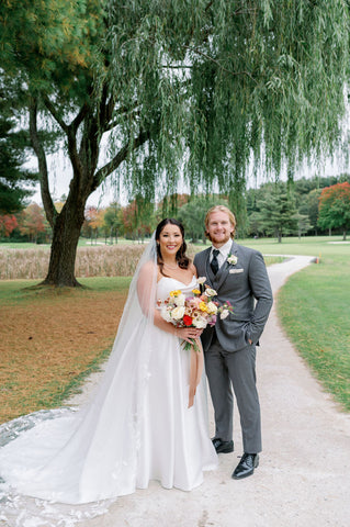 Imae og bride and groom during couples portraits at the Cape Club of Sharon, a Massachusetts wedding venue. They are under a beautiful old tree. The groom is in a grey suit ith black tie. The bride is wearing a white satin gown holding a lush bridal bouquet in a muted fall color palette with dahlias, marigolds, sedum, celosia, ranunculus, lisianthus, mums, leucadendron, asclepia, eucalyptus, and amaranthus.