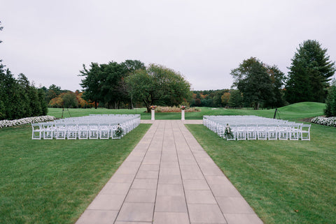 Image of outdoor ceremony space at the Cape Club of Sharon, a Massachuetts wedding venue. The image shows the long walkway used as an aisle with white graden chairs on either side, over lush green grass.