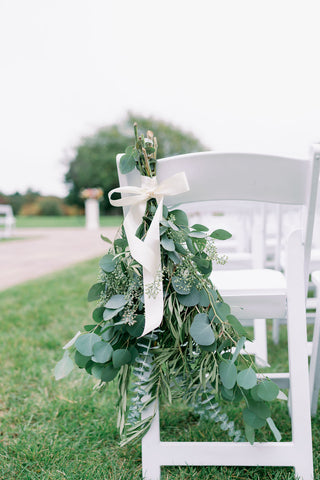 Image of greenery floral design on wedding ceremony chairs. The image shows gathered greenery tied up on the back of a white garden ceremony chair.