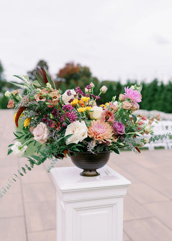 Image of muted fall wedding ceremony flowers on a white pedestal at the Cape Club of Sharon. a Massachusetts wedding venue. The flowers include dahlias, marigolds, sedum, celosia, ranunculus, lisianthus, mums, leucadendron, asclepia, eucalyptus, and amaranthus.