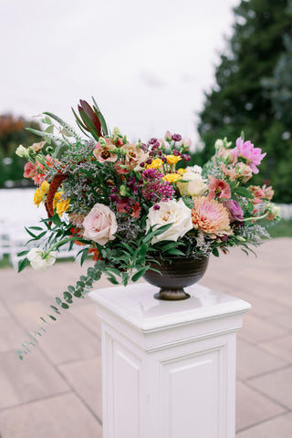 Image of muted fall wedding ceremony flowers on a white pedestal at the Cape Club of Sharon, a Massachusetts wedding venue. Flowers include dahlias, marigolds, sedum, celosia, ranunculus, lisianthus, mums, leucadendron, asclepia, eucalyptus, and amaranthus.