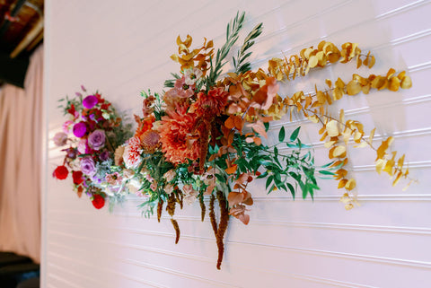 Image of the wall in the reception space at the Cape Club of Sharon, a Massachusetts wedding venue. The photo zooms in on the wall that has a stunning statement floral installation hanging on the white wall. The color palette is muted fall colors. The flowers include dahlias, marigolds, sedum, celosia, ranunculus, lisianthus, mums, leucadendron, asclepia, eucalyptus, and amaranthus.