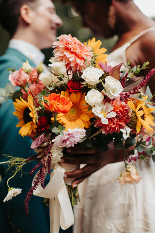 Photo of a couple on their wedding day. The bridal bouquet is the focal point with the couple out of focus behind. The bouquet is bright and joyful with sunflowers and flowers in shades of yellow, orange, coral and white. 