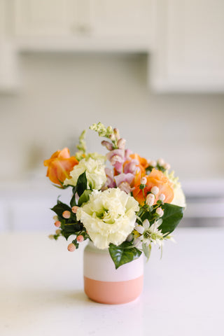 Image of small floral arrangement in a pink and white vase. The flowers include shades of white and orange.