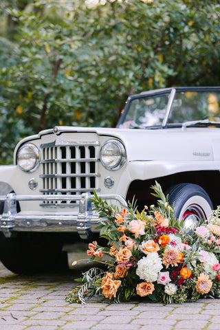 Image of vintage white car next to statement wedding floral design. The vibrant flower design includes roses, hydrangeas, raununculus, dahlias and more in shades of orange.