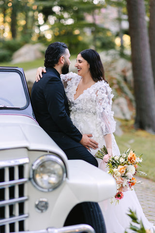 Image of bride and groom in front of a vintage car. The bride is holding a vibrant hued bouquet in shades of orange and early fall colors.