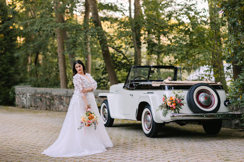 Image of a bride standing next to a vintage wedding rental car from Timless Wheel Events located in Massachusetts. The bride is carrying a vibrant wedding bouquet in shades of orange featuring roses and raununculus.