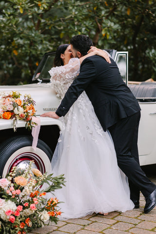 Image of bride and groom in front of a vintage car. The bride is in a long sleeved dress. The event looks elegant between the vintage white car and luxe florals in vibrant hues.