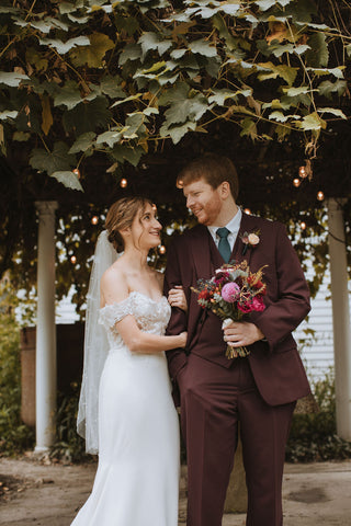 Bride and groom wedding portrait at the Herb Lyceum after their outdoor ceremony. Husband is holding the brides bouquet which has deep, rich, vibrant fall colors including deep reds, vibrant pink and pale pink with greenery.