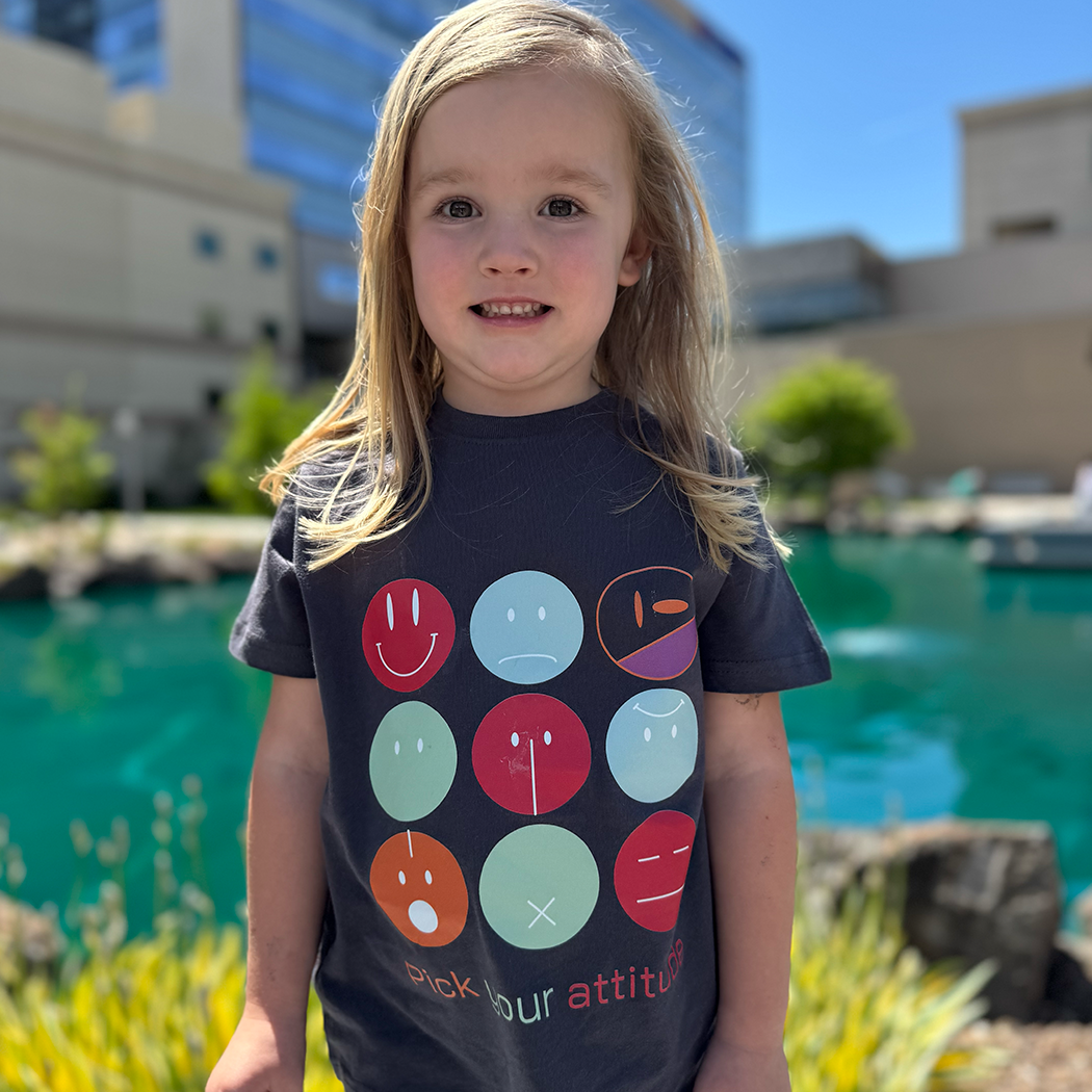 A young child with blonde hair smiles while standing outdoors in front of a pond and modern buildings. The child is wearing a dark gray graphic apparel t-shirt with a grid of colorful smiley faces and the text "Pick your attitude" printed on it, along with light pink leggings and gray shoes.
