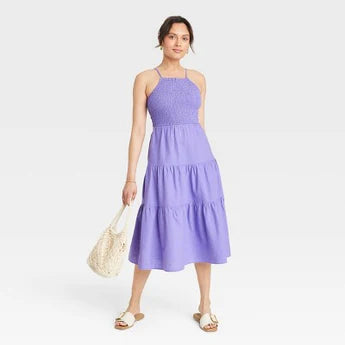 Woman wearing a sleeveless purple dress, holding a woven bag.