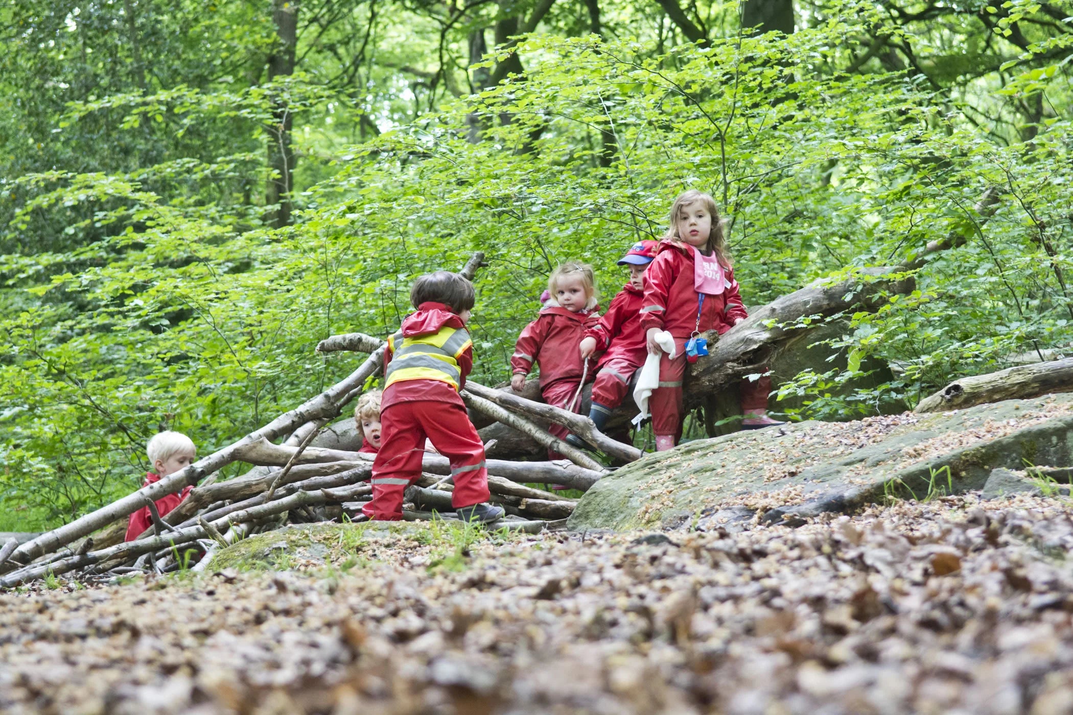 Children playing in the woods, wearing red rain suits near a pile of branches.