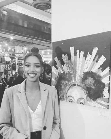 Black woman in tan coat, white top and black jeans smiles next to her artwork displayed in an art gallery.