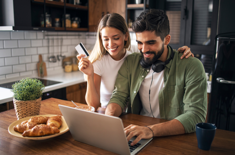 Man and women in kitchen siting down at table with laptop open and card in hand smiling