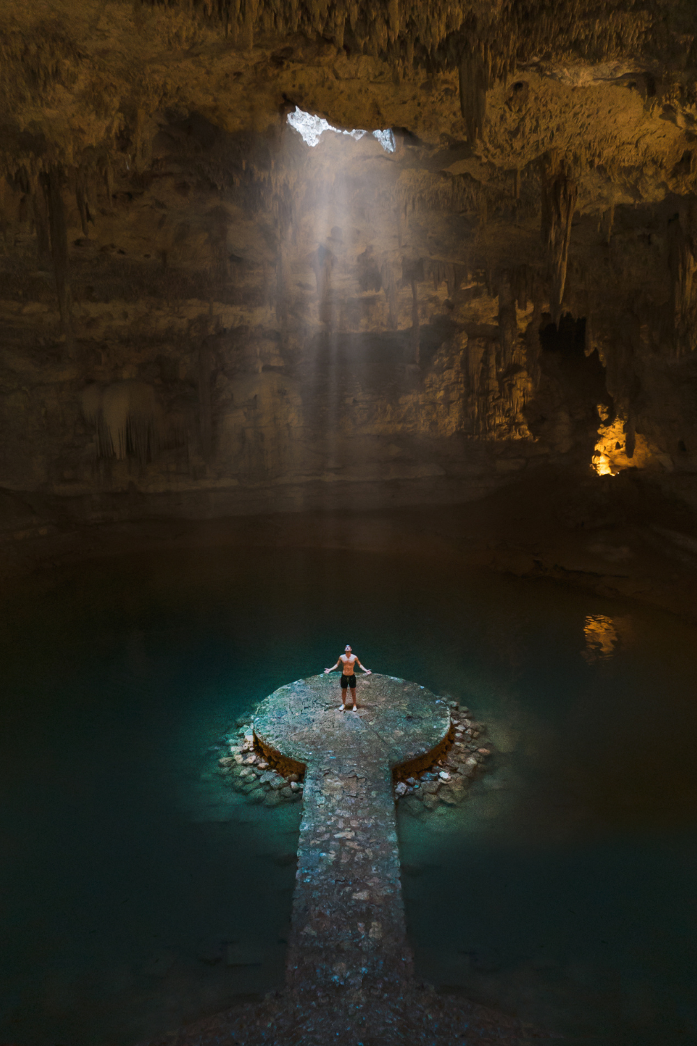 Man standing on stone landing in large cave under opening with water all around and light shinning through