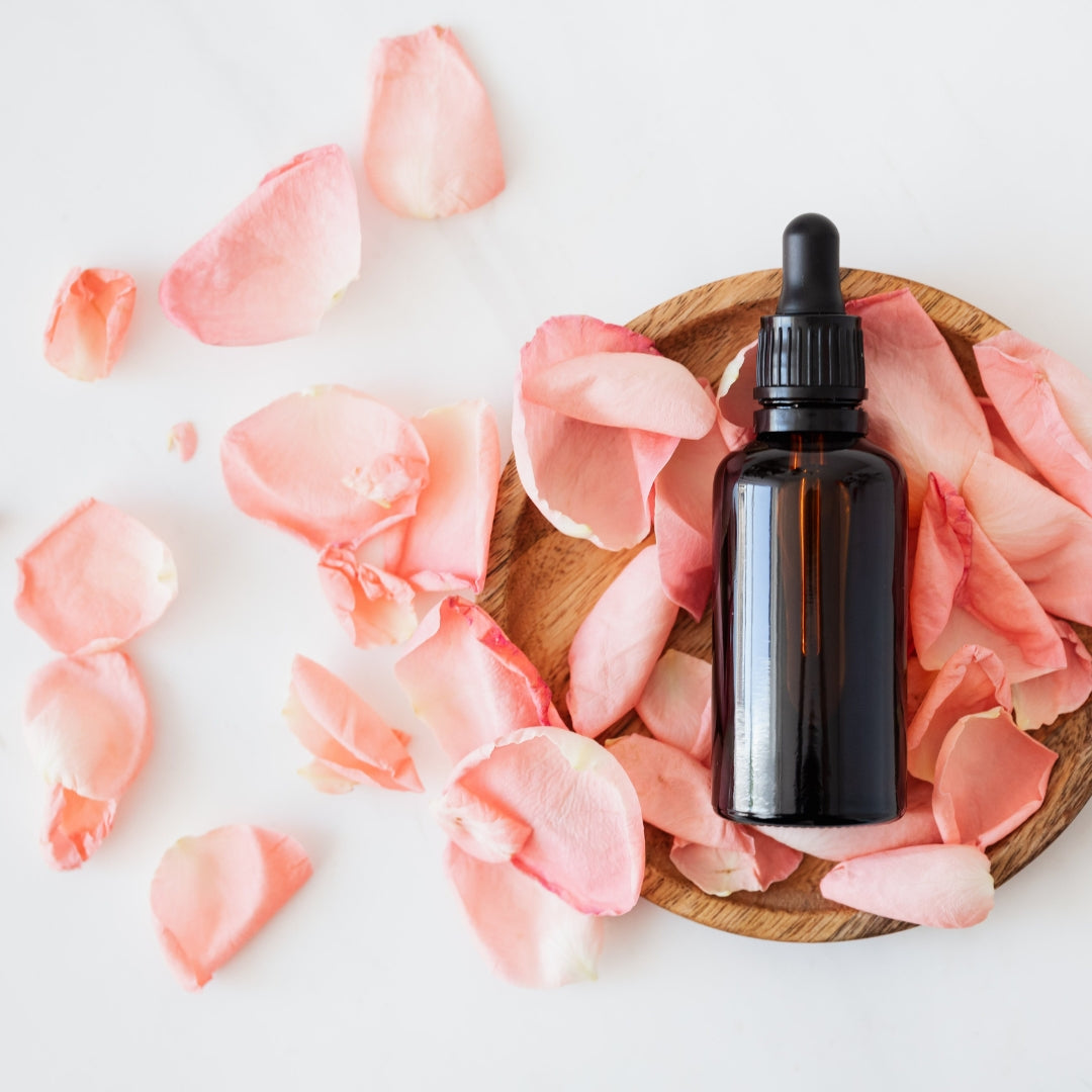 This image shows an essential oil dropper bottle on a wooden plate surrounded by pink rose petals on a white background.