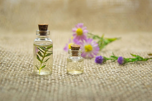 This image features two clear glass bottles with cork lids, one containing green twigs with purple flowers on a textured beige background.