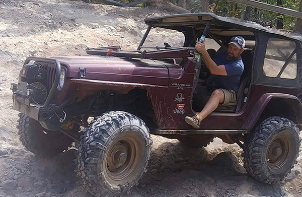 A man drives a maroon Jeep on a dirt road.