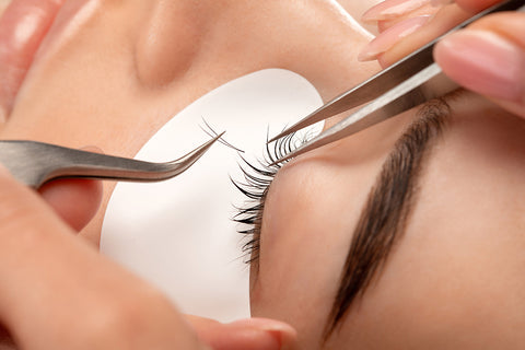 closeup look of a professional technician applying eyelash extensions to a woman's natural lashes using tweezers, carefully attaching a 2D fan to one real lash