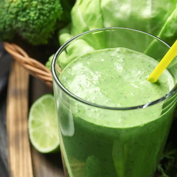 Ein Glas frisch gepresster Brokkolisaft mit einem gelben Strohhalm, einer halben Limette, Brokkoli und einem Kopf Salat als Dekoration auf einem Holztisch.