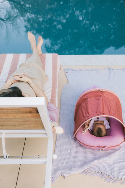 woman resting on a sun lounger whilst her baby is lounging in a DockATot Deluxe+ dock with Cabana Kit