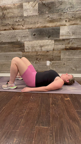 A person in pink shorts and black tank top laying on yoga mat with feet on floor and knees up